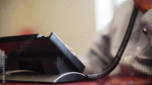 A close up reverse angle from behind a landline office phone that has it's handset picked up and a phone number is dialed on i'ts keypad, by male hands in warm light.