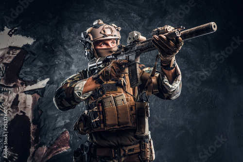 Elite unit  special forces soldier in camouflage uniform holding an assault rifle with a laser sight and aims at the target. Studio photo against a dark textured wall