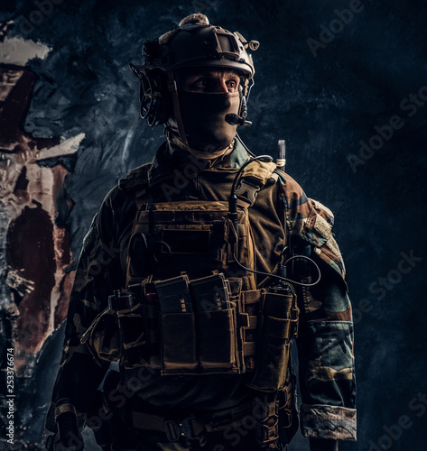 Private security service contractor in camouflage uniform and helmet with walkie-talkie. Studio photo against a dark textured wall
