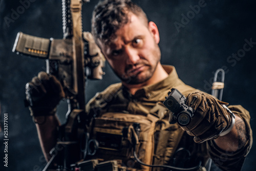 Special forces soldier wearing body armor holding assault rifle and aiming a gun at the camera. Studio photo against a dark textured wall