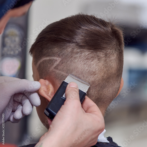 ber making a haircut to a cute European boy using cutting machine. He is making hair design.