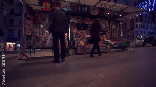 Low rotating pan shot across people in retail area of Madrid at night exploring tourist kiosk stall on sidewalk illuminated by decorative lights & traffic.