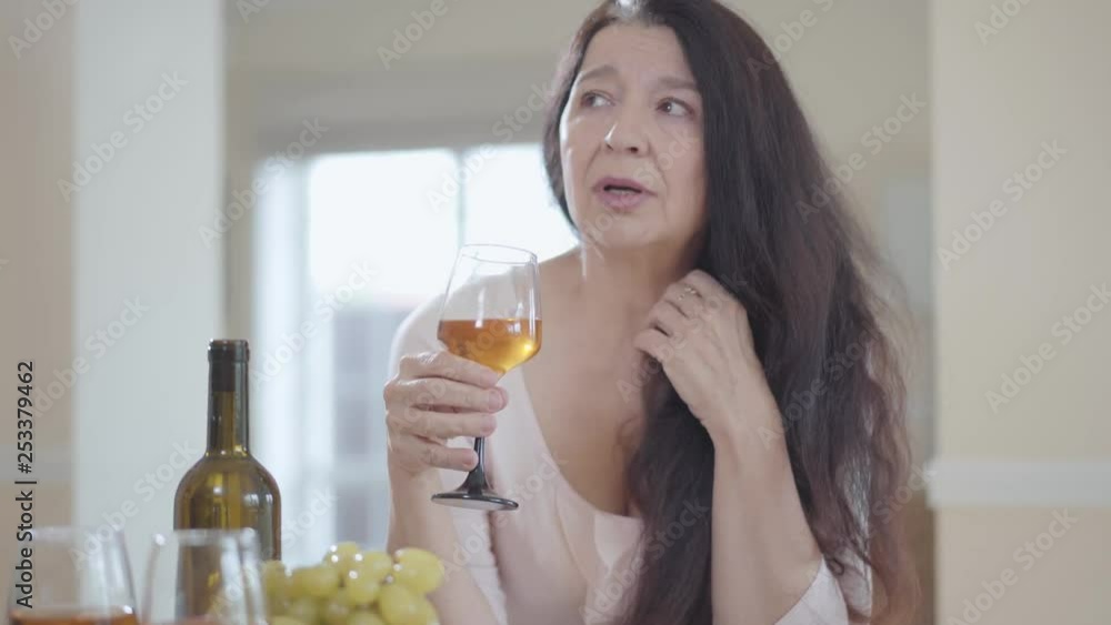 Portrait of sad senior woman with gorgeous long dark hair leaned on the table indoors close up. Lady is crying, she takes glass and going to drink wine. Portrait of sad senior woman with gorgeous long dark hair leaned on the table indoors close up. Lady is crying, she takes glass and going to drink wine.