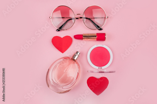 Valentine's day flatlay with beauty products: bottle of perfumes, red lipstick, rose blush, gradient round sunglasses, women heands and paper heart isolated on pink background. photo