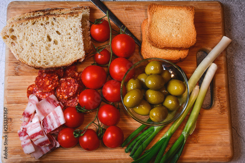 The homemade white bread on a wooden board with organic bacon, cut sausage, cherry tomato, onions and green olives.