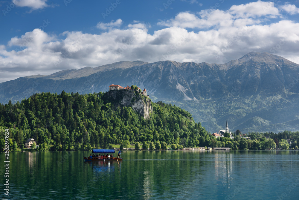 Traditional Pletna boat rowing tourists on Lake Bled with Bled castle on cliff and St Martin church Sol massive of Karawanks mountains Slovenia