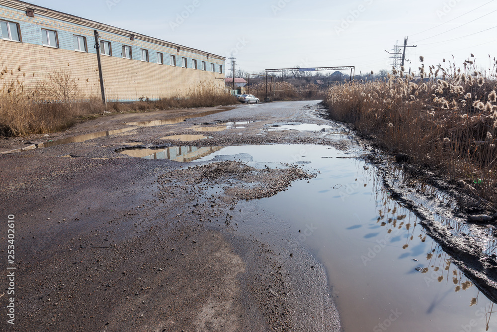  Damaged asphalt road with potholes caused by freezing and thawing cycles during the winter. Bad road with cars in water pits