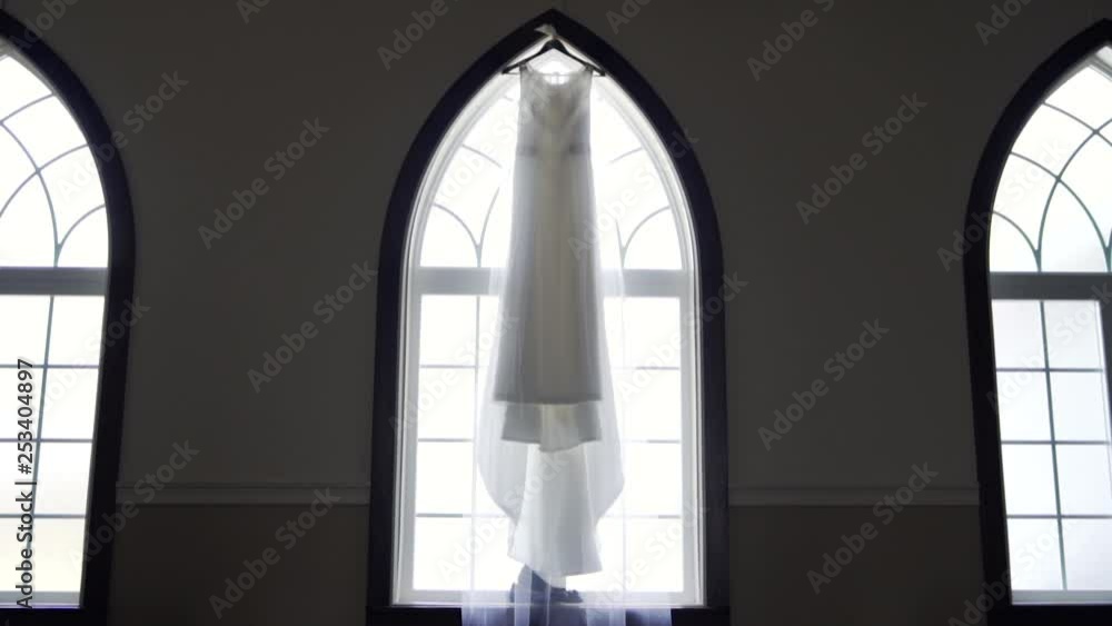 Wide vista of a bride's beautiful wedding dress hanging from a large window sill in an old church building - Slow push in slider shot Wide vista of a bride's beautiful wedding dress hanging from a large window sill in an old church building - Slow push in slider shot