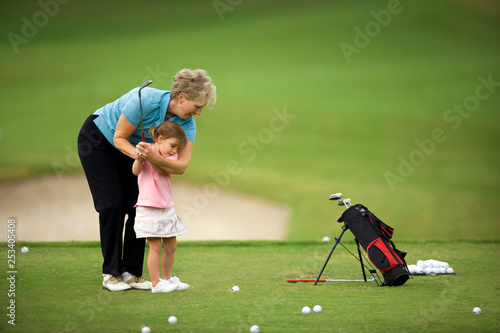 Senior woman teaching her young granddaughter how to swing a golf club on a golf course.