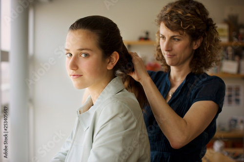 Portrait of a teenage girl getting her hair fixed by her mother inside their home.