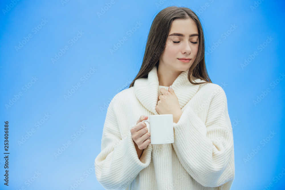 Interestingly, a young woman in a knitted white sweater. Holds a white cup of coffee or tea in his hands. Isolated against the background of a blue wall, studio portrait. Concept of a way of life of Interestingly, a young woman in a knitted white sweater. Holds a white cup of coffee or tea in his hands. Isolated against the background of a blue wall, studio portrait. Concept of a way of life of