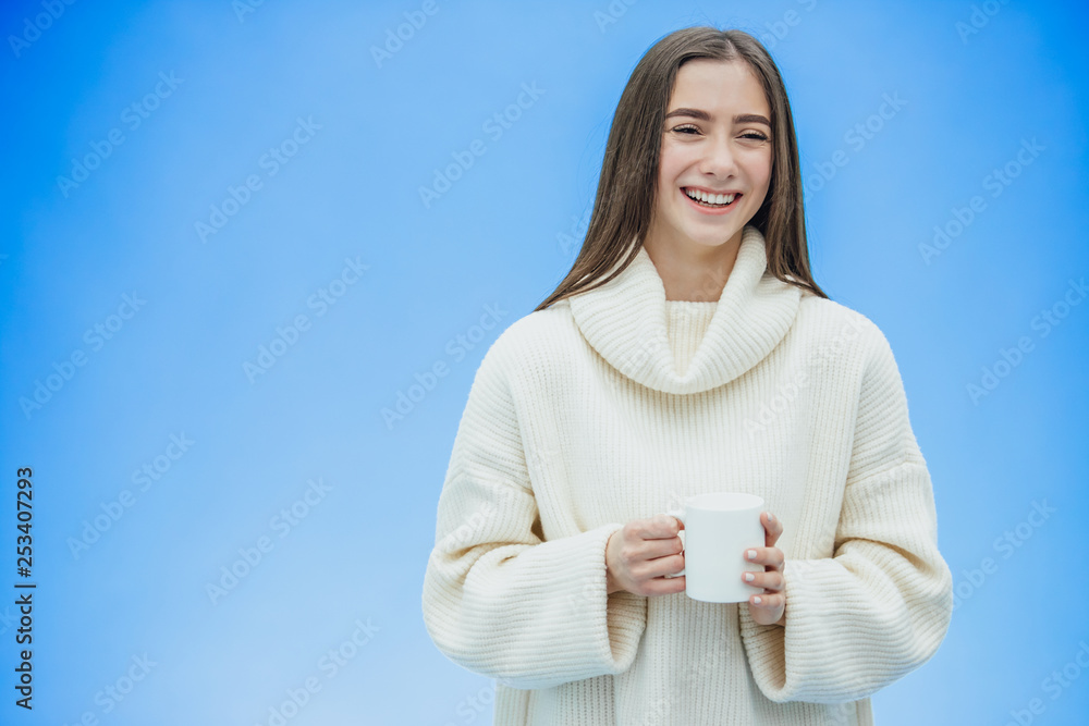 Interestingly, a young woman in a knitted white sweater. Holds a white cup of coffee or tea in his hands. Isolated against the background of a blue wall, studio portrait. Concept of a way of life of Interestingly, a young woman in a knitted white sweater. Holds a white cup of coffee or tea in his hands. Isolated against the background of a blue wall, studio portrait. Concept of a way of life of