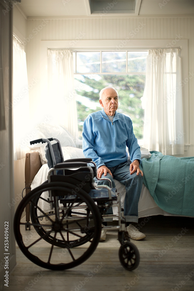 Contemplative senior man sitting on the edge of his bed next to a wheelchair. Contemplative senior man sitting on the edge of his bed next to a wheelchair.