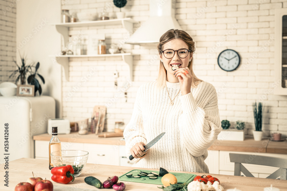 Joyful good-looking lady biting piece of cucumber in kitchen