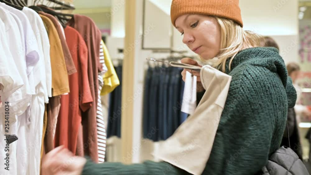 A girl in a green sweater and a yellow hat walks through a store of things and chooses what to buy. Touches things on hangers and looks at price tags