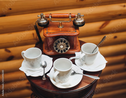 An old, antique, rarity telephone with a disk dialing set is standing on the table near empty white cups on a wooden background. Cloce up. photo