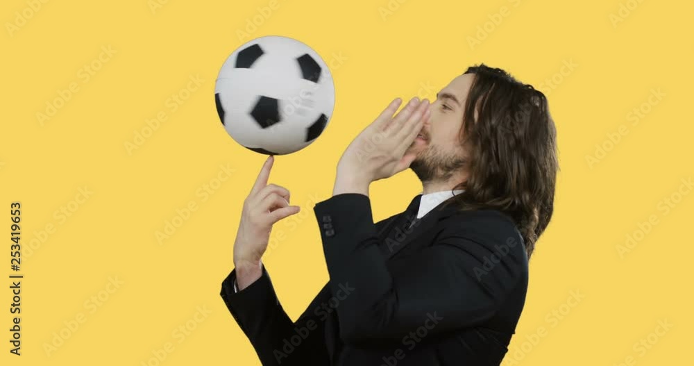 Caucasian attractive young man in the suit and tie standing on the yellow background and spinning football ball on his finger. Caucasian attractive young man in the suit and tie standing on the yellow background and spinning football ball on his finger.