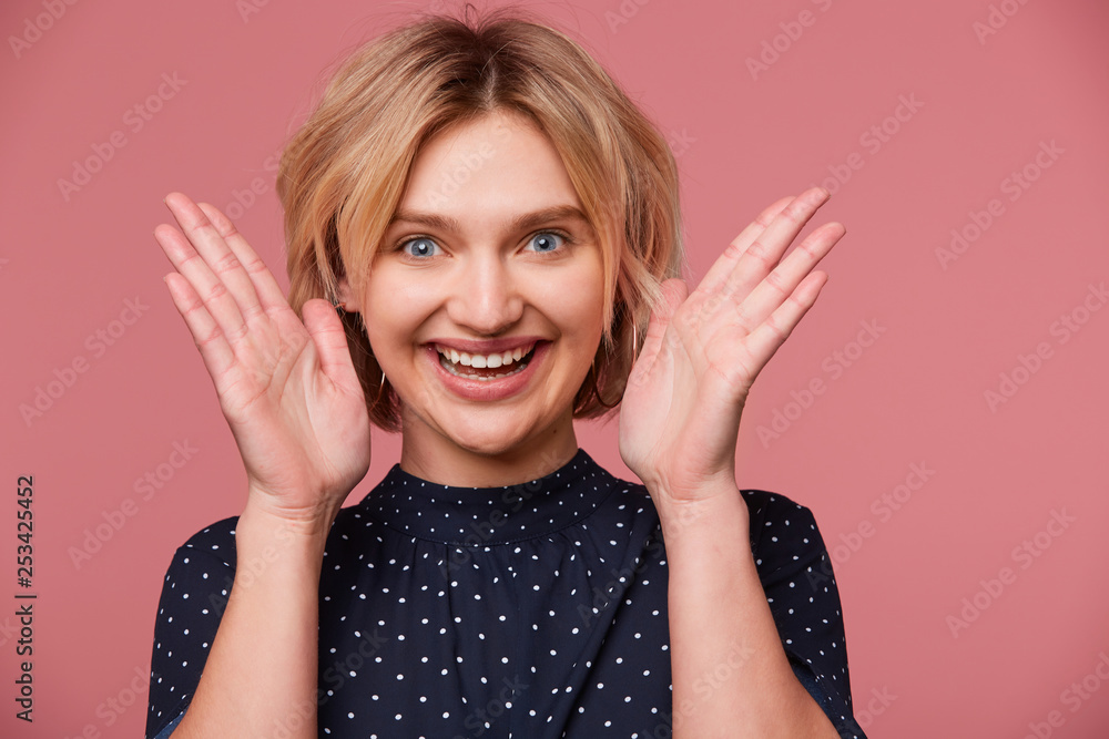 Close up of excited young beautiful attractive blonde woman dressed in blouse with polka dots, keeps palms near the face has exited face expression, showing positive, smiling, happy, over pink wall Close up of excited young beautiful attractive blonde woman dressed in blouse with polka dots, keeps palms near the face has exited face expression, showing positive, smiling, happy, over pink wall