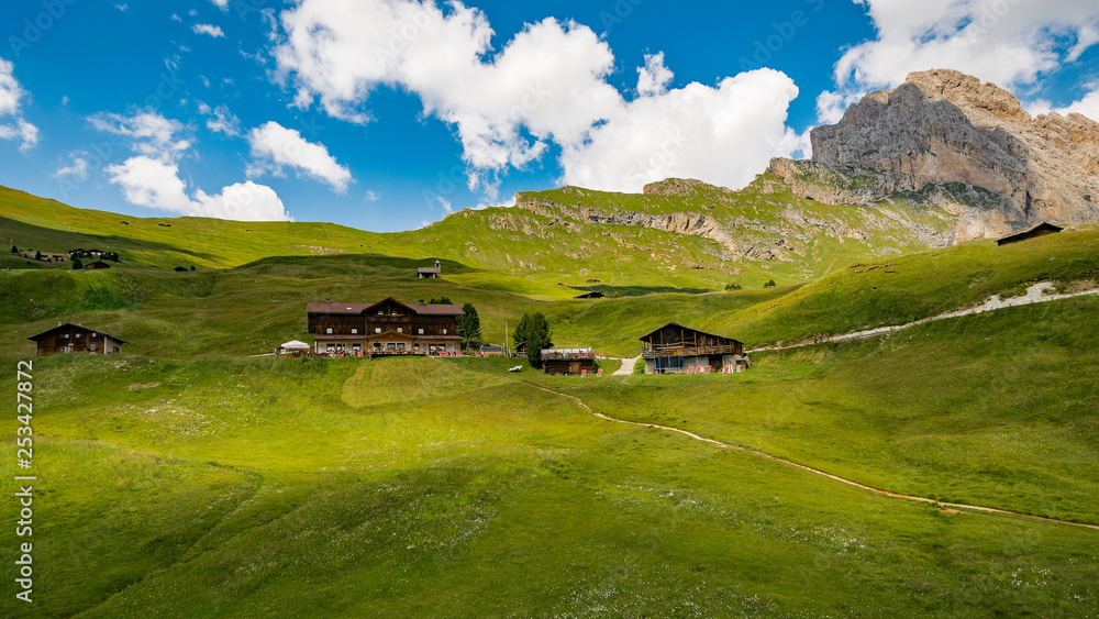 Group of beautiful mountain huts on a gentle slope of the Dolomites with a rocky rocky peak in the background. Blue sky with big white clouds.