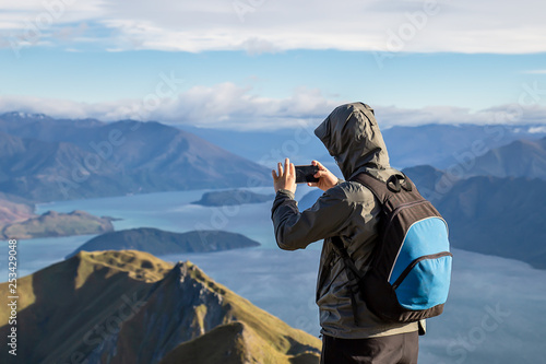 Young man on top of hill taking photo on cellphone smartphone of beautiful lake and mountain landscape in New Zealand