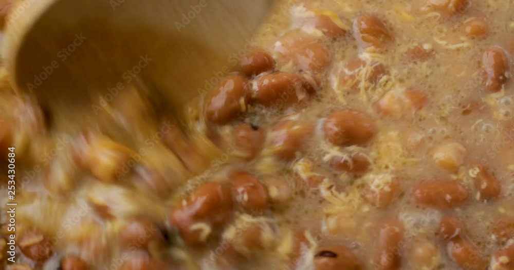 Close video of stirring melted shredded cheddar cheese in fava beans and broth while heating with a wood spoon illuminated with natural lighting. Close video of stirring melted shredded cheddar cheese in fava beans and broth while heating with a wood spoon illuminated with natural lighting.
