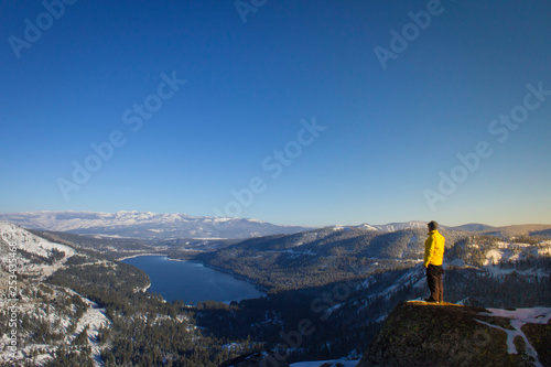 A man in a yellow jacket looks down on a blue lake photo