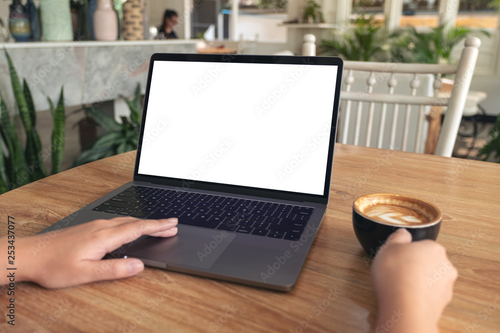 Mockup image of woman's hands typing and touching on laptop touchpad with blank white desktop screen while drinking coffee on wooden table in cafe Mockup image of woman's hands typing and touching on laptop touchpad with blank white desktop screen while drinking coffee on wooden table in cafe