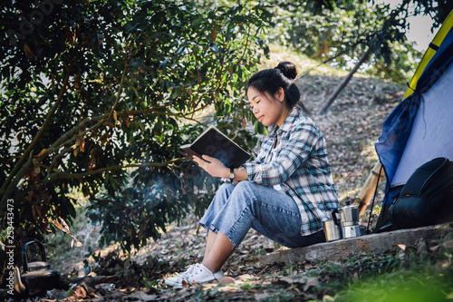 Asian young woman in tartan shirt sitting is reading a book in outside the tent. Alone camping in forest.