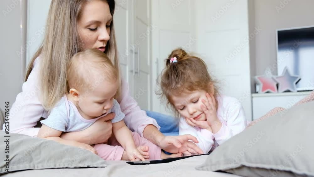 Positive family mother, daughter and son are cheerfully watching a tablet with children's films sitting in a cozy bedroom. Concept of care and education for children Positive family mother, daughter and son are cheerfully watching a tablet with children's films sitting in a cozy bedroom. Concept of care and education for children