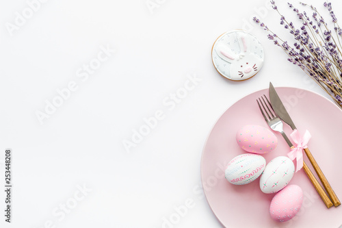 Easter dining table in pastel colors. Plate, cutlery, painted eggs, gingerbread and lavender branches on white background top view copy space