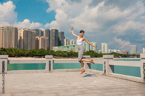 Cheerful and beautiful girl having fun on vacation, jumping on the pier on background of city. In China, Hainan city of Sanya. Town Sanya is popular tourist destination in China. View of island in bay © timofeev