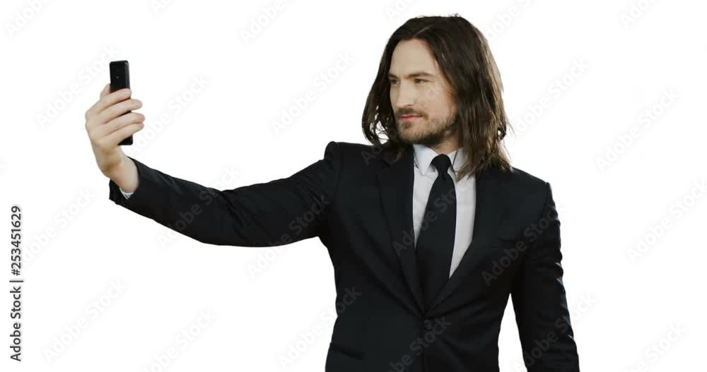 Portrait shot of the attractive young businessman in the suit and tie making nice and serious selfies on the smartphone while standing in front of the camera on the white wall background. Portrait shot of the attractive young businessman in the suit and tie making nice and serious selfies on the smartphone while standing in front of the camera on the white wall background.