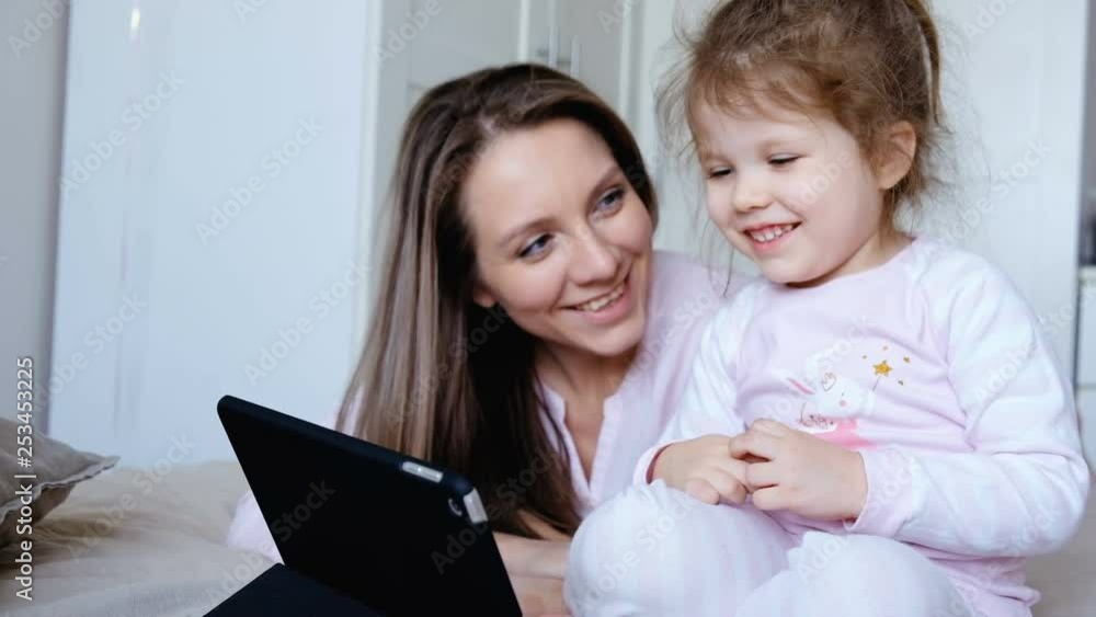 Positive little girl and her mom together watching a funny cartoon on a tablet lying on a sofa in a cozy room. Concept of affection for each other and care Positive little girl and her mom together watching a funny cartoon on a tablet lying on a sofa in a cozy room. Concept of affection for each other and care