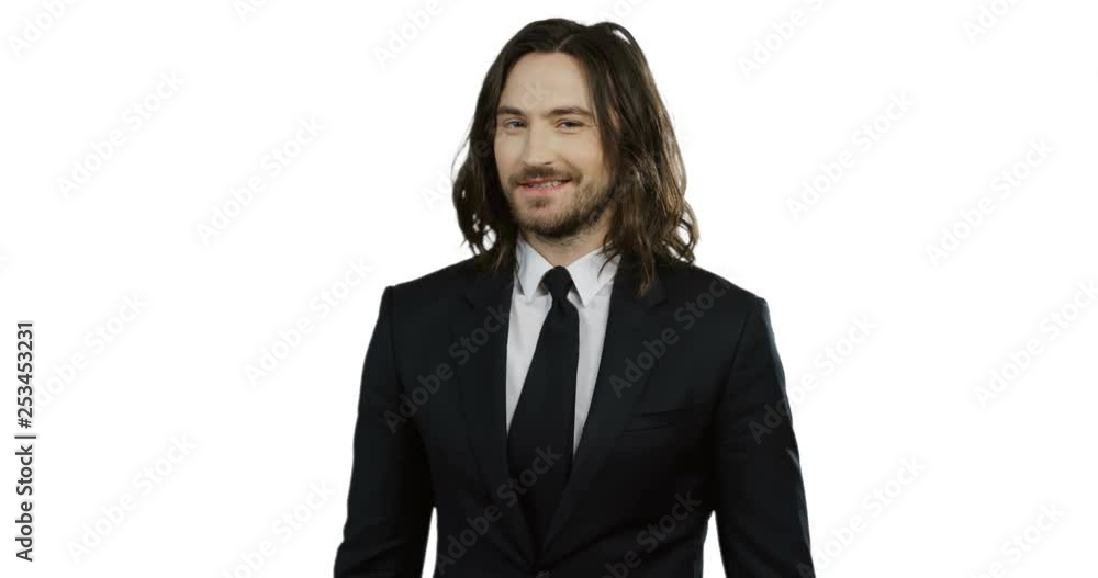 Portrait shot of the handsome young Caucasian man with long hair in the suit and tie looking in the camera and smiling while standing on the white screen background. Portrait shot of the handsome young Caucasian man with long hair in the suit and tie looking in the camera and smiling while standing on the white screen background.