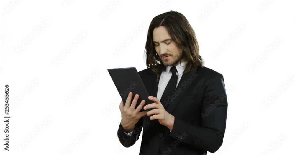 Portrait shot of the brunette young serious man in the black suit and tie scrolling and taping on the tablet on the white wall background. Portrait shot of the brunette young serious man in the black suit and tie scrolling and taping on the tablet on the white wall background.