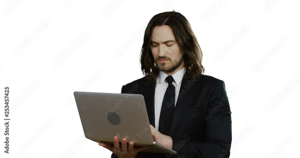 Caucasian young businessman in the black suit and tie scrolling and taping on the laptop computer, then looking in the camera seriously on the white background. Caucasian young businessman in the black suit and tie scrolling and taping on the laptop computer, then looking in the camera seriously on the white background.