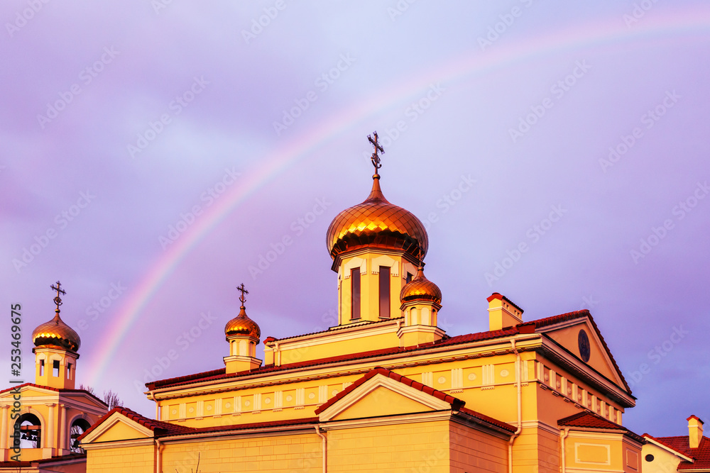 The golden domes of the Orthodox Christian church in the rays of the evening sun setting against the background of a bright multi-colored rainbow after the rain. Rainbow, Good News, Gospel