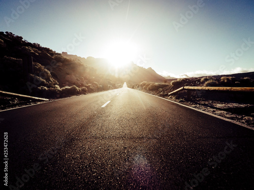 Long way road at the mountain with sun in front and sunlight effect - ground point of view with black asphalt and white lines - driving and travel concept