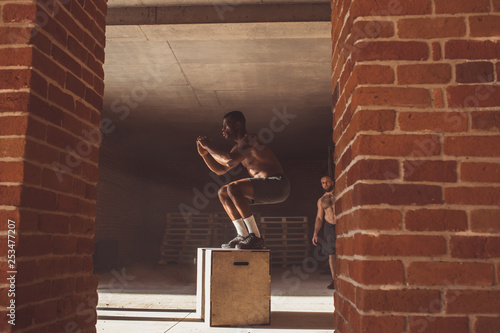 Handsome strong african man with bare torso jumping onto a plyobox in a cross training gym with big panoramic windows on background photo