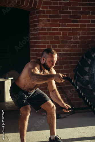 Handsome muscular bare chested gym man is doing battle rope exercise while working out at cross-fit training.