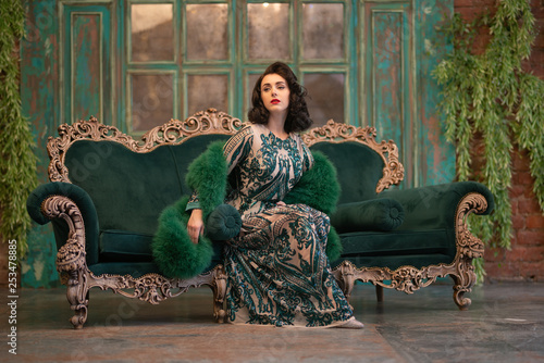 elegant caucasian girl in a luxurious long sequins lace dress with a green fluffy boa in her hands posing in the retro room with vintage luxury furniture