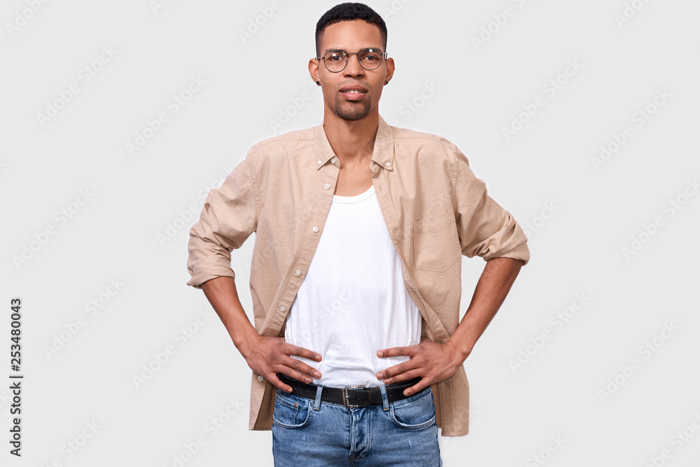 Fototapeta premium Studio portrait of young African American man wearing casual white t-shirt and beige shirt, looking to the camera through round eyeglasses and posing over white background. People emotion concept