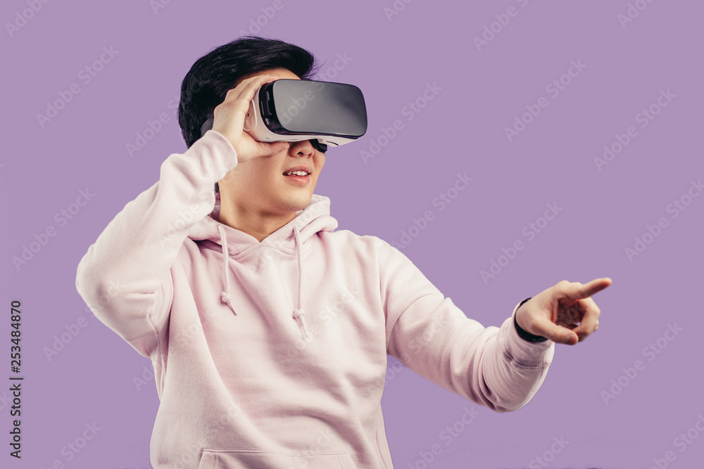 Korean young man surprised by something in virtual reality, wearing virtual reality glasses showing gesture isolated on studio shot on violet wall background Korean young man surprised by something in virtual reality, wearing virtual reality glasses showing gesture isolated on studio shot on violet wall background
