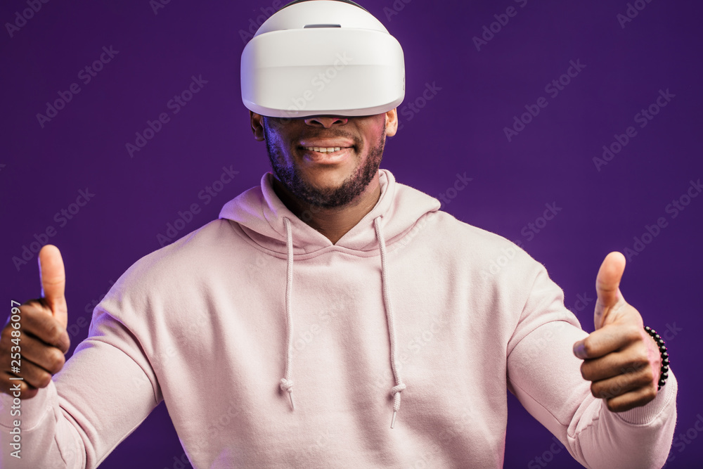 African American guy in white hoodie is playing a virtual reality game using head-mounted display, feeling excitement and joy, isolated over purple background with copyspace