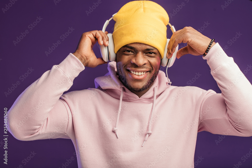 Young African man in hoodie, having the fashion sense of a street urchin, turned a bit in headphones isolated on violet background , holding earphones with hands listening to favorite tracks Young African man in hoodie, having the fashion sense of a street urchin, turned a bit in headphones isolated on violet background , holding earphones with hands listening to favorite tracks