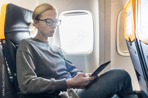Blonde casual caucasian woman wearing glasses reading on digital e-reader while traveling by airplane. Commercial transportation by planes.