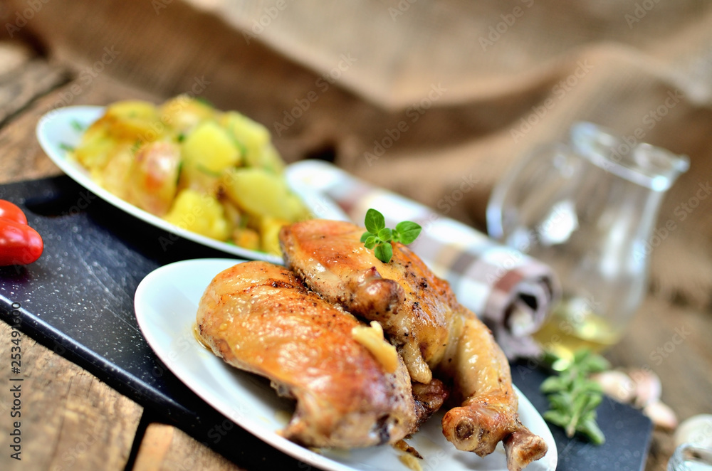 Golden grilled chicken legs on a plate and roasted potatoes, rosemary, jug with oil, oregano, salt, pepper and garlic on a wooden table Golden grilled chicken legs on a plate and roasted potatoes, rosemary, jug with oil, oregano, salt, pepper and garlic on a wooden table