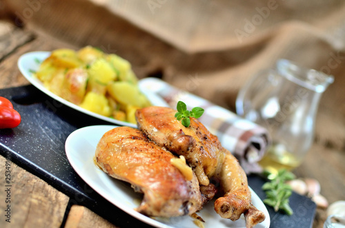 Golden grilled chicken legs on a plate and roasted potatoes, rosemary, jug with oil, oregano, salt, pepper and garlic on a wooden table