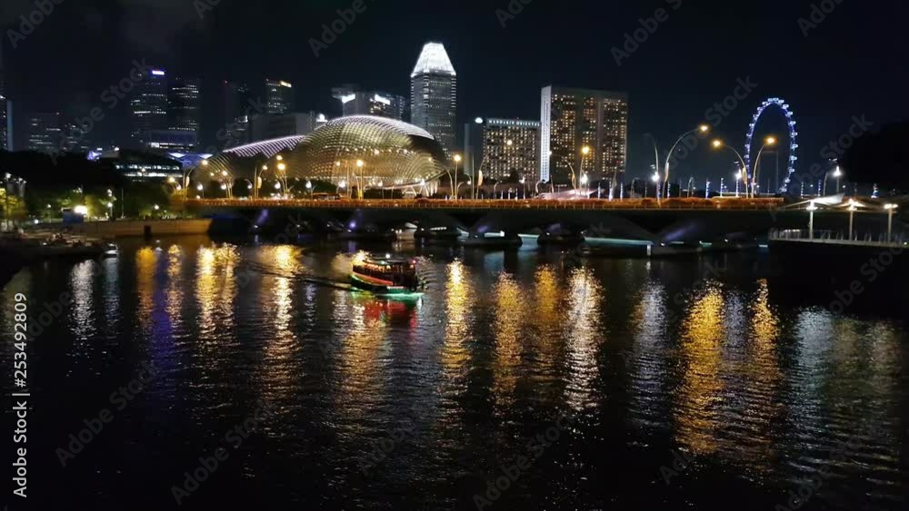 View of Singapore skyline at night along a Singapore River with a bumboat moving View of Singapore skyline at night along a Singapore River with a bumboat moving