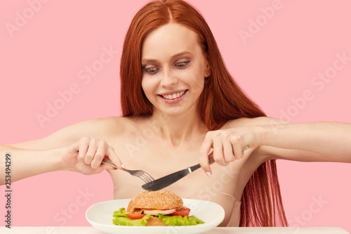 Excited red-haired skinny half naked woman looking unhealthy with greedy eyes being ready to eat burger  sitting at table isolated over pink background. Eating disorder.